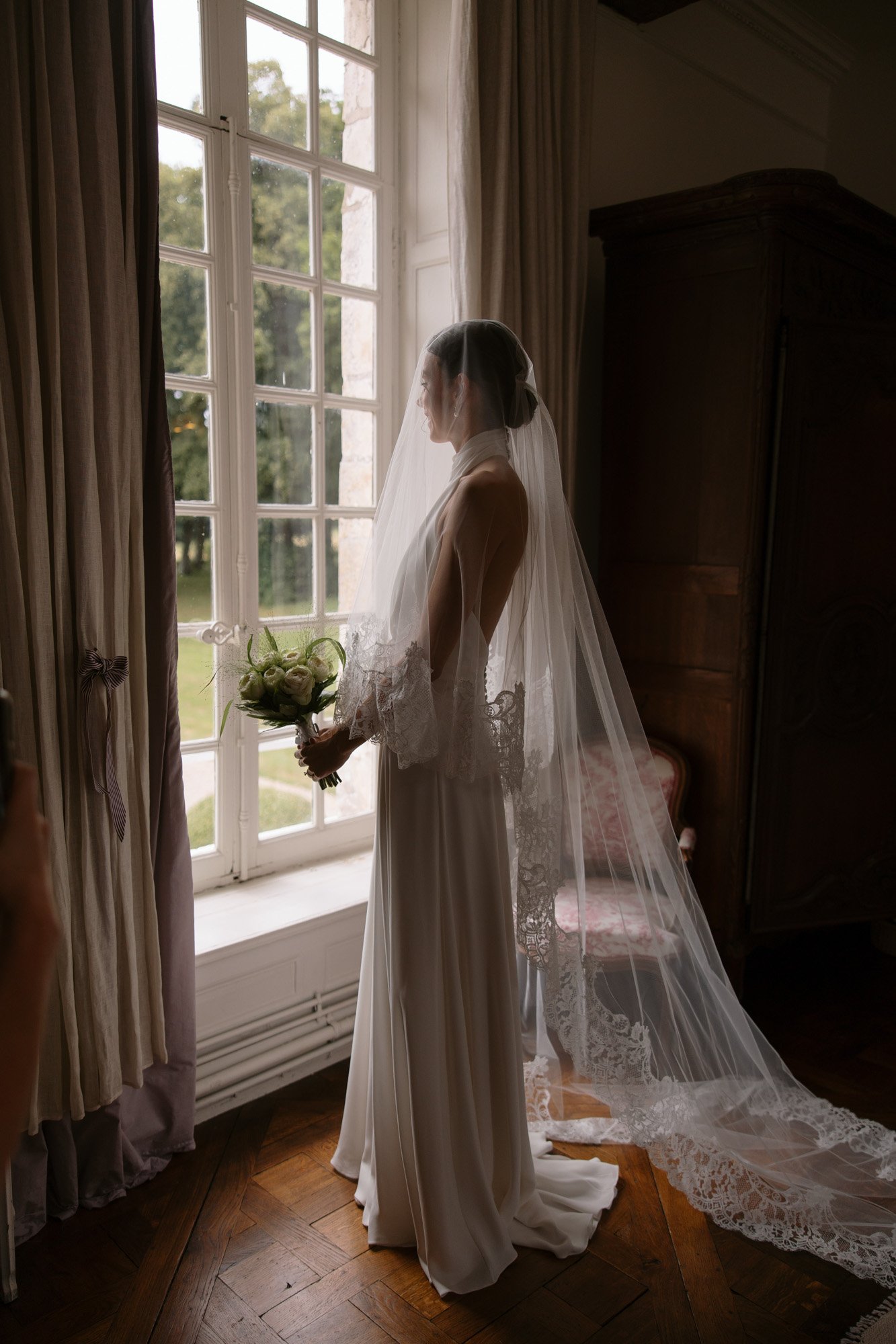 A bride in a white wedding dress and veil stands by a large window, holding a bouquet and looking outside in a softly lit room. Captured by Northern France wedding photographer.