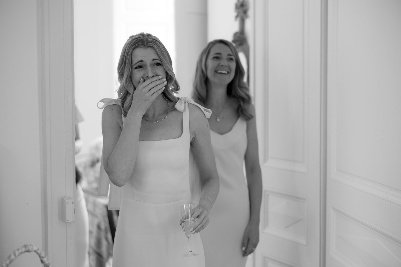Two women in white dresses stand indoors; one holds a glass and covers her mouth with her hand, appearing emotional, while the other smiles in the background.