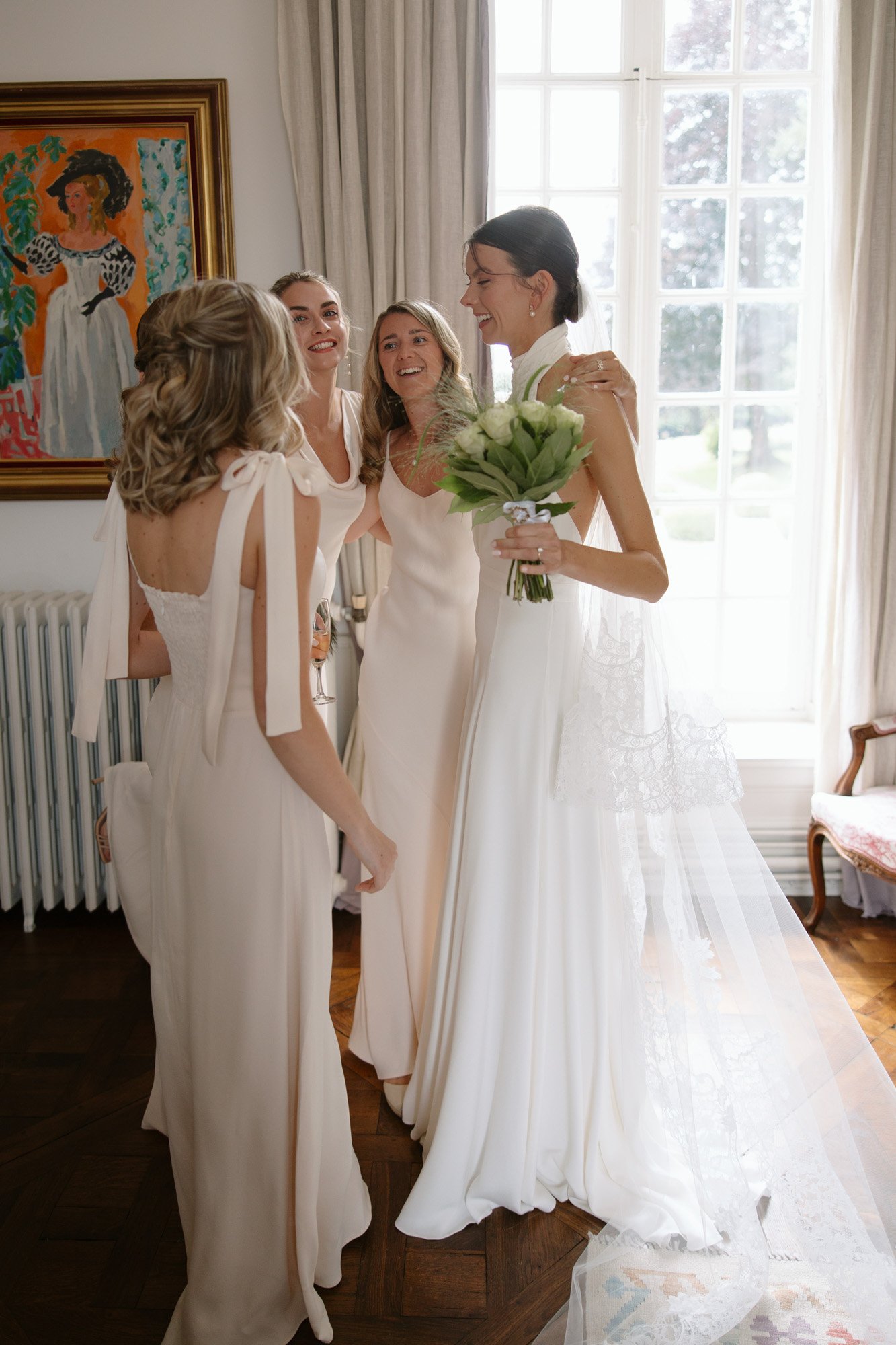 Four women in light-colored dresses stand together indoors, smiling and talking. One holds a bouquet and wears a veil, suggesting she is the bride. Large window and painting in background.