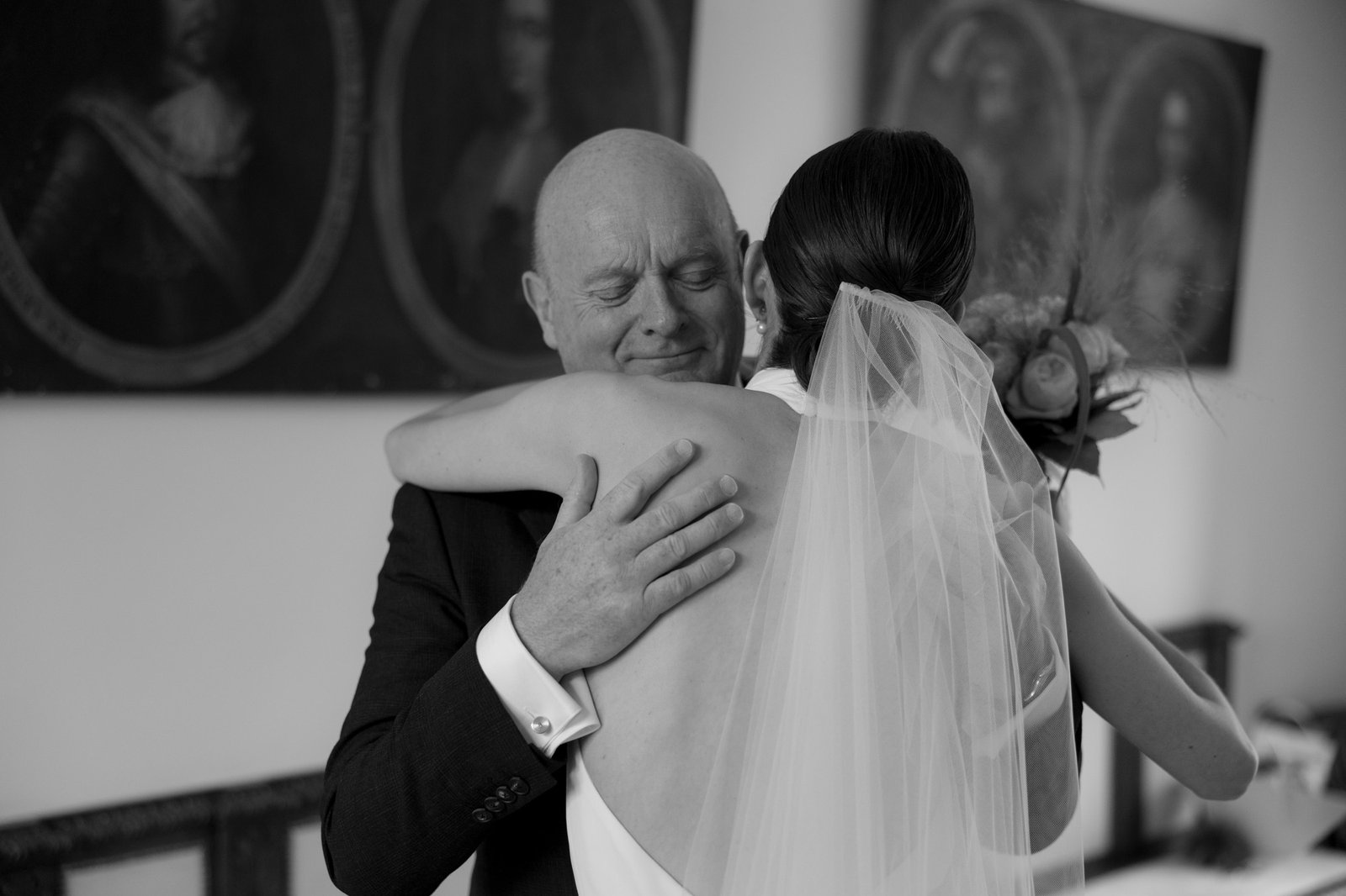 A bald man in a suit hugs a woman in a wedding dress and veil indoors; he appears emotional with eyes closed.