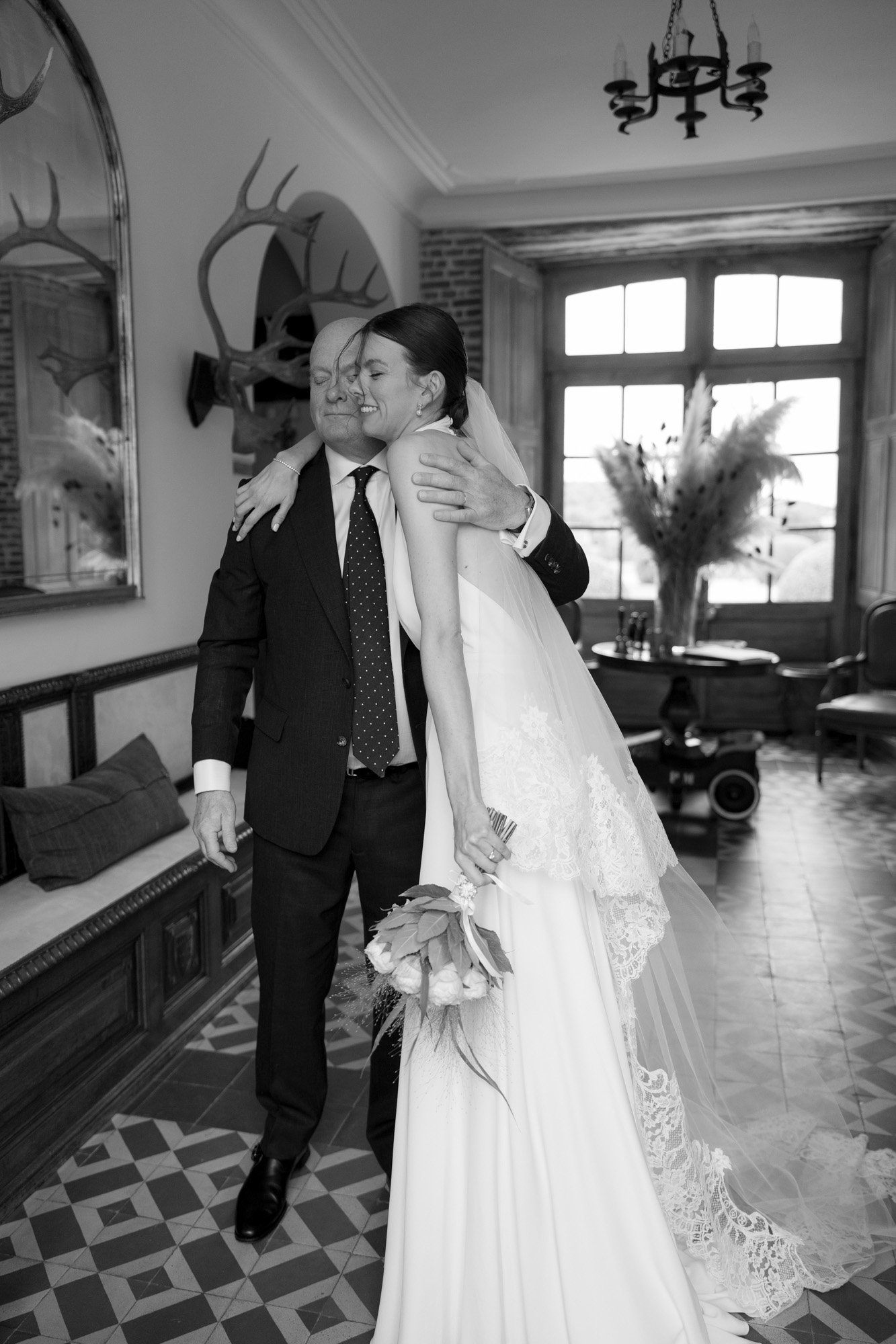 A bride in a wedding dress smiles and hugs a man in a suit indoors, holding a bouquet, with a window and decorative antlers in the background. Captured by Northern France wedding photographer.