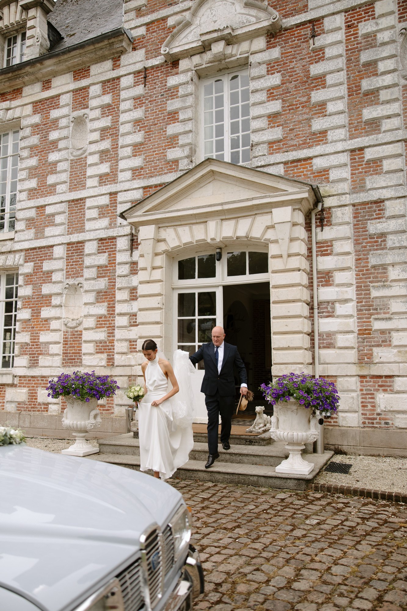 A bride and an older man walk down the steps of a brick building with flowers in large planters, approaching a white car parked on a cobblestone driveway.
