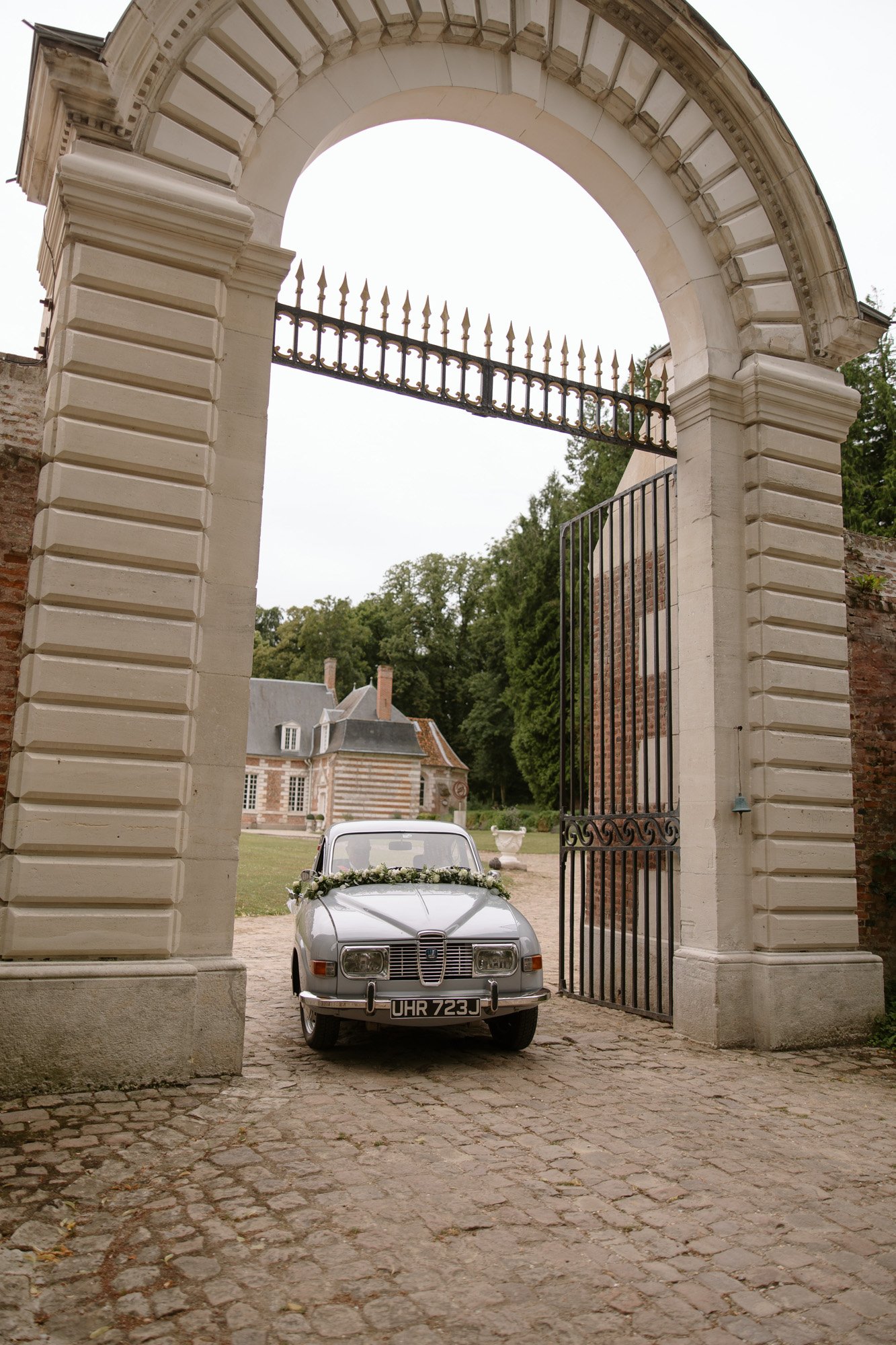 A vintage silver car parked under a large stone archway with an ornate gate, with a historic building and trees visible in the background.