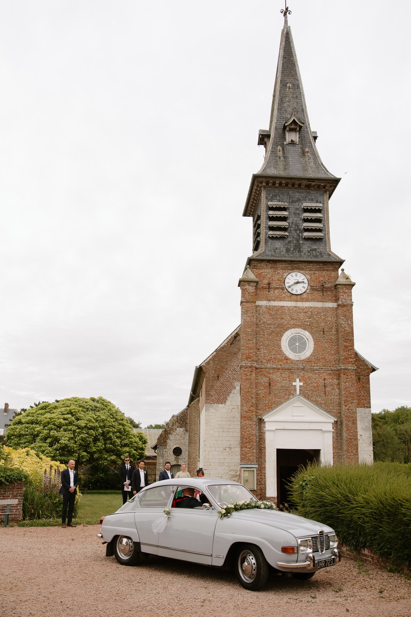 A vintage silver car decorated with flowers is parked in front of a brick church with a tall steeple, while a few people stand nearby. Captured by Northern France wedding photographer.