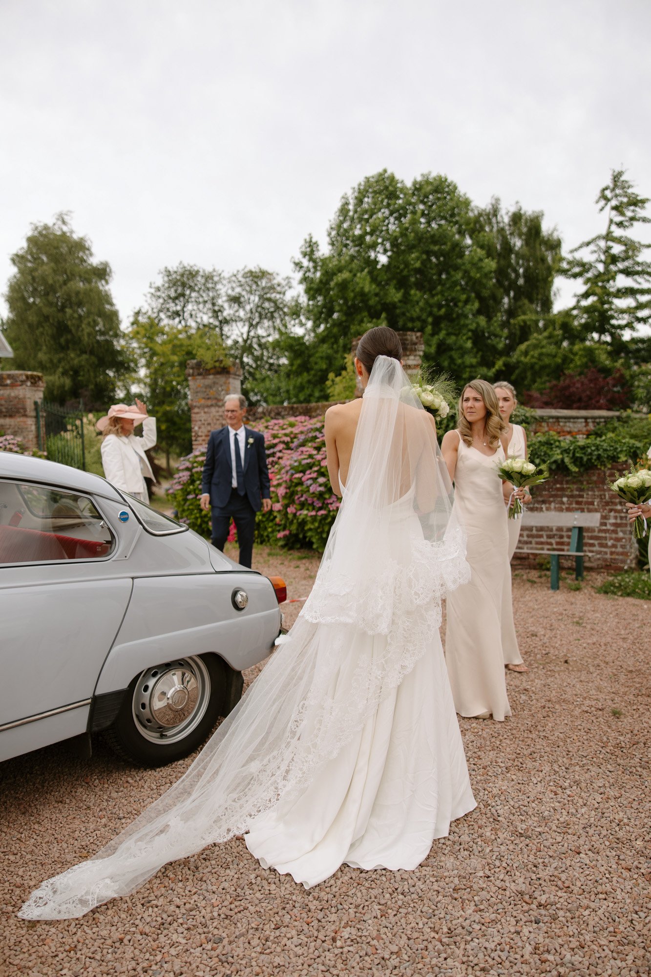 A bride in a white dress and veil stands near a vintage car, with wedding guests and bridesmaids in light dresses gathered outside in a garden setting.