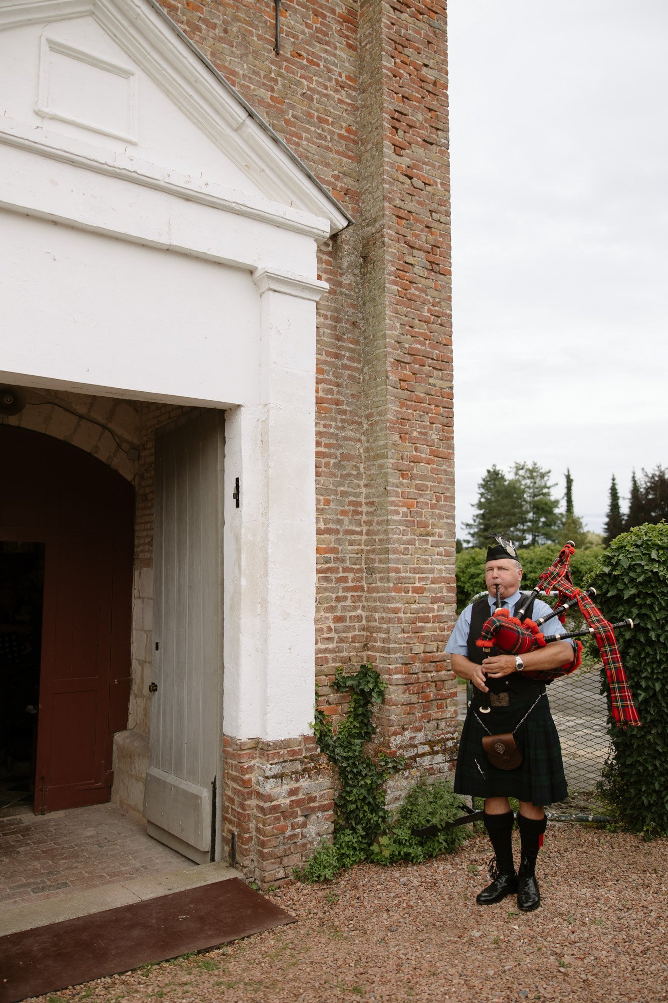 A man in traditional Scottish attire plays bagpipes outside a brick building with a white entrance.