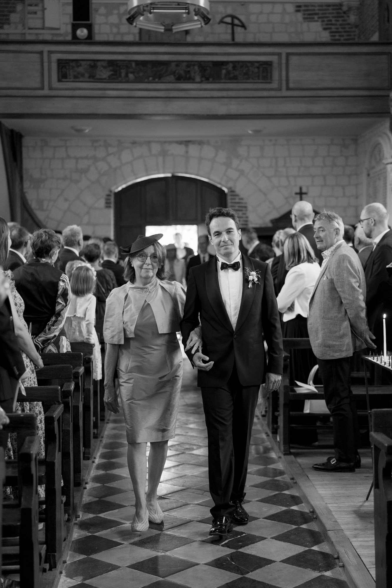 A formally dressed man and an older woman walk arm in arm down the aisle of a church, surrounded by seated guests.