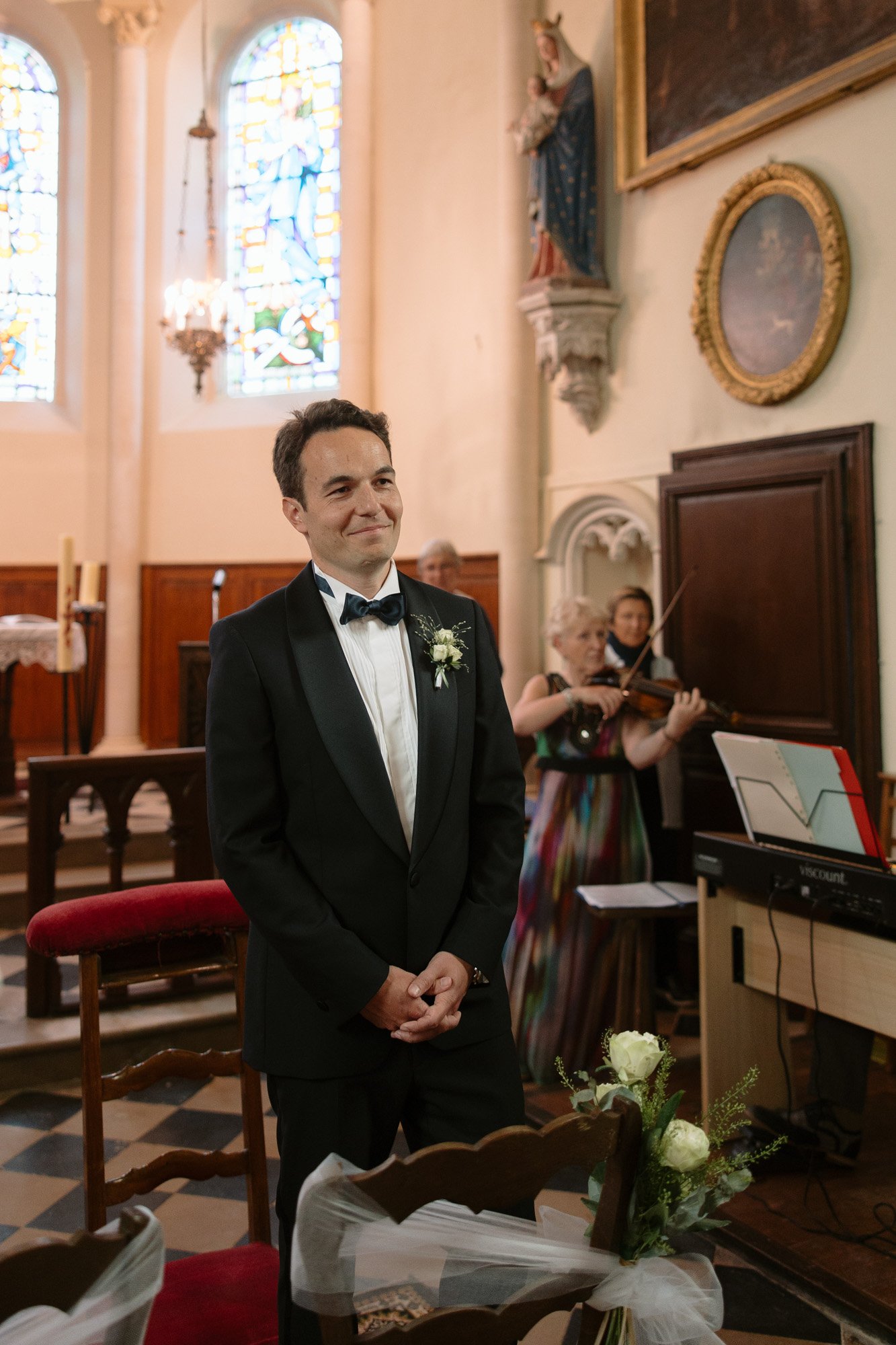 A man in a black tuxedo stands and smiles in a church, with stained glass windows behind and a violinist playing nearby.