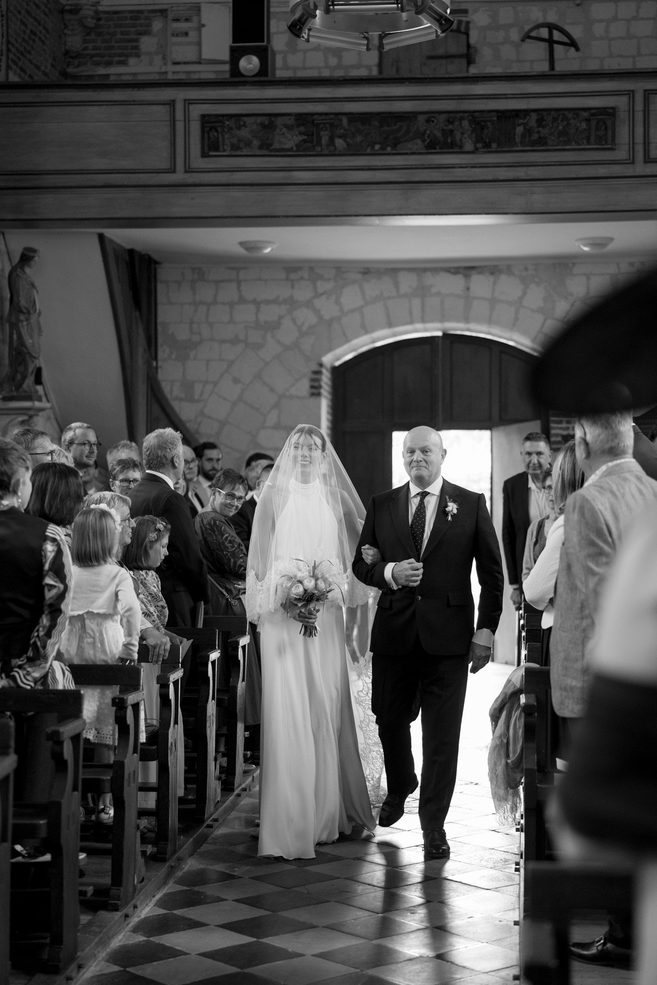 A bride in a wedding dress and veil walks down the aisle with an older man in a suit inside a church, surrounded by seated guests. Captured by Northern France wedding photographer.