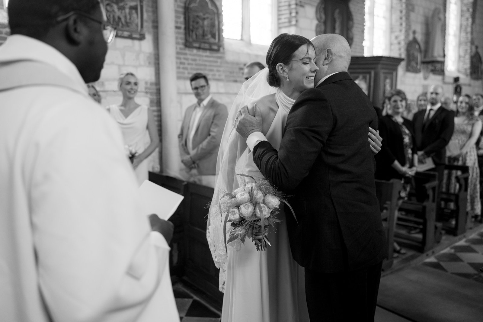 A bride hugs an older man, likely her father, in a church during a wedding ceremony, with guests and an officiant present.