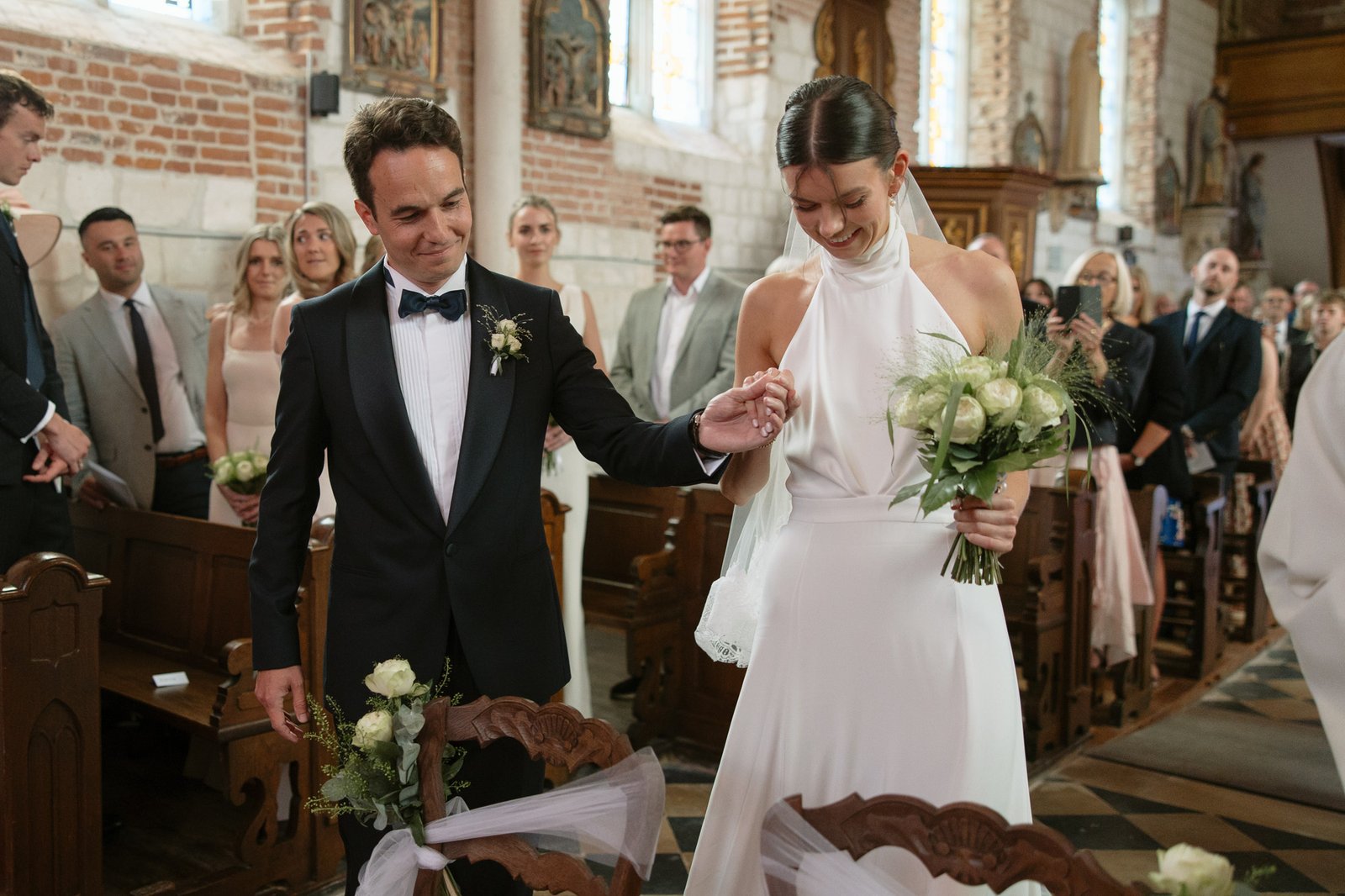 A bride and groom stand together at the front of a church, holding hands and flowers, surrounded by seated and standing guests.