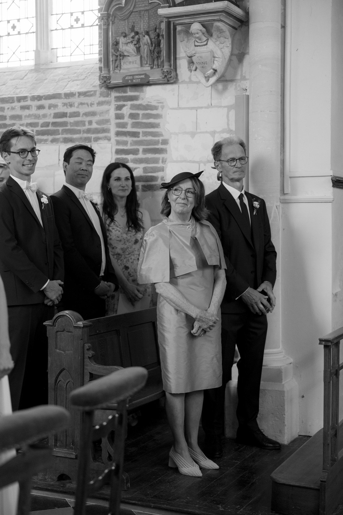 A group of formally dressed people stand and smile inside a church, with wooden pews and brick walls visible in the background.
