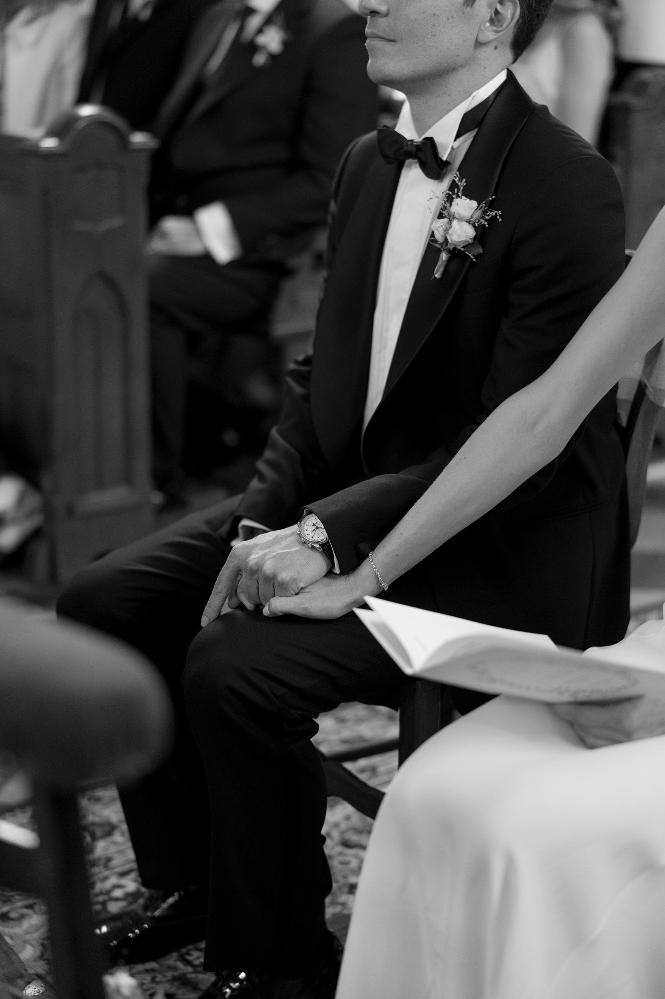 A couple dressed in formal attire sits side by side in a church, holding hands during a ceremony.