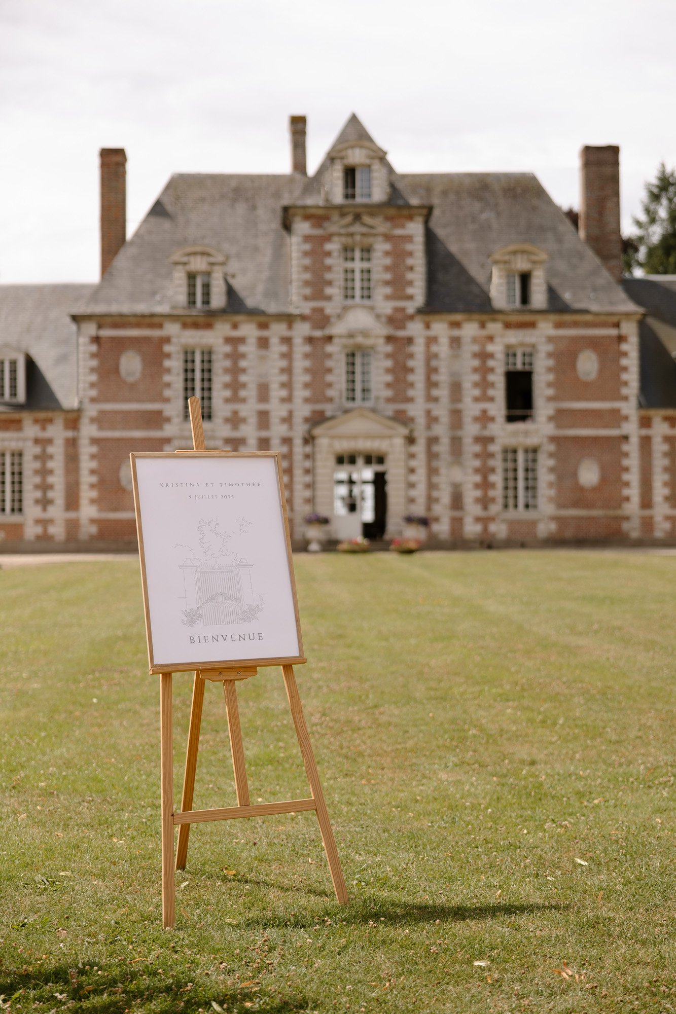 A welcome sign on an easel stands on a lawn in front of a large, historic brick and stone building with dormer windows.