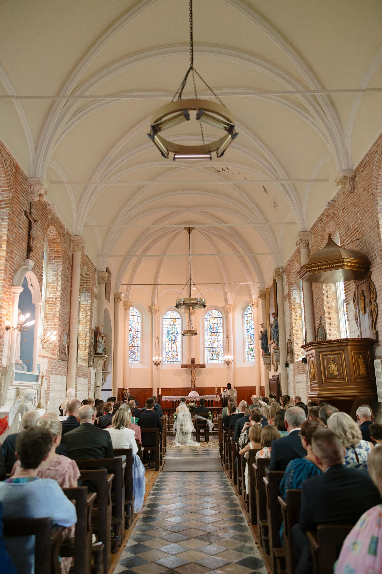 Bride and groom sit at the front of a church during a wedding ceremony, surrounded by seated guests facing the altar and stained glass windows.