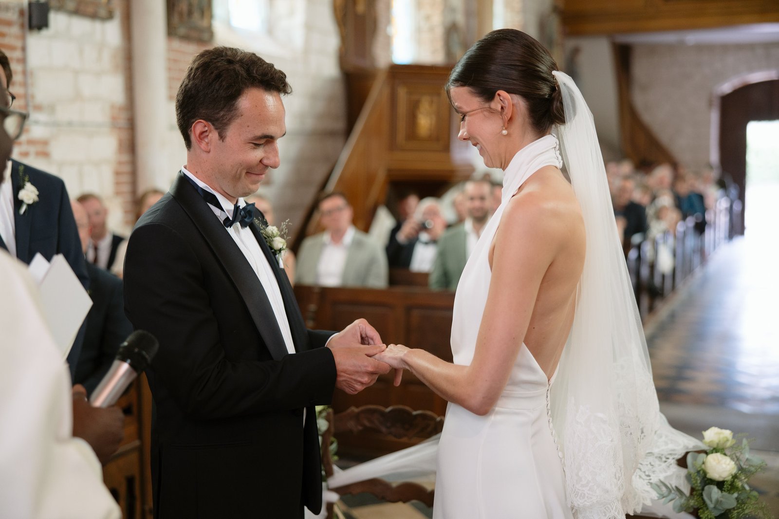 A bride and groom exchange rings during their wedding ceremony inside a church, with guests seated in the background.