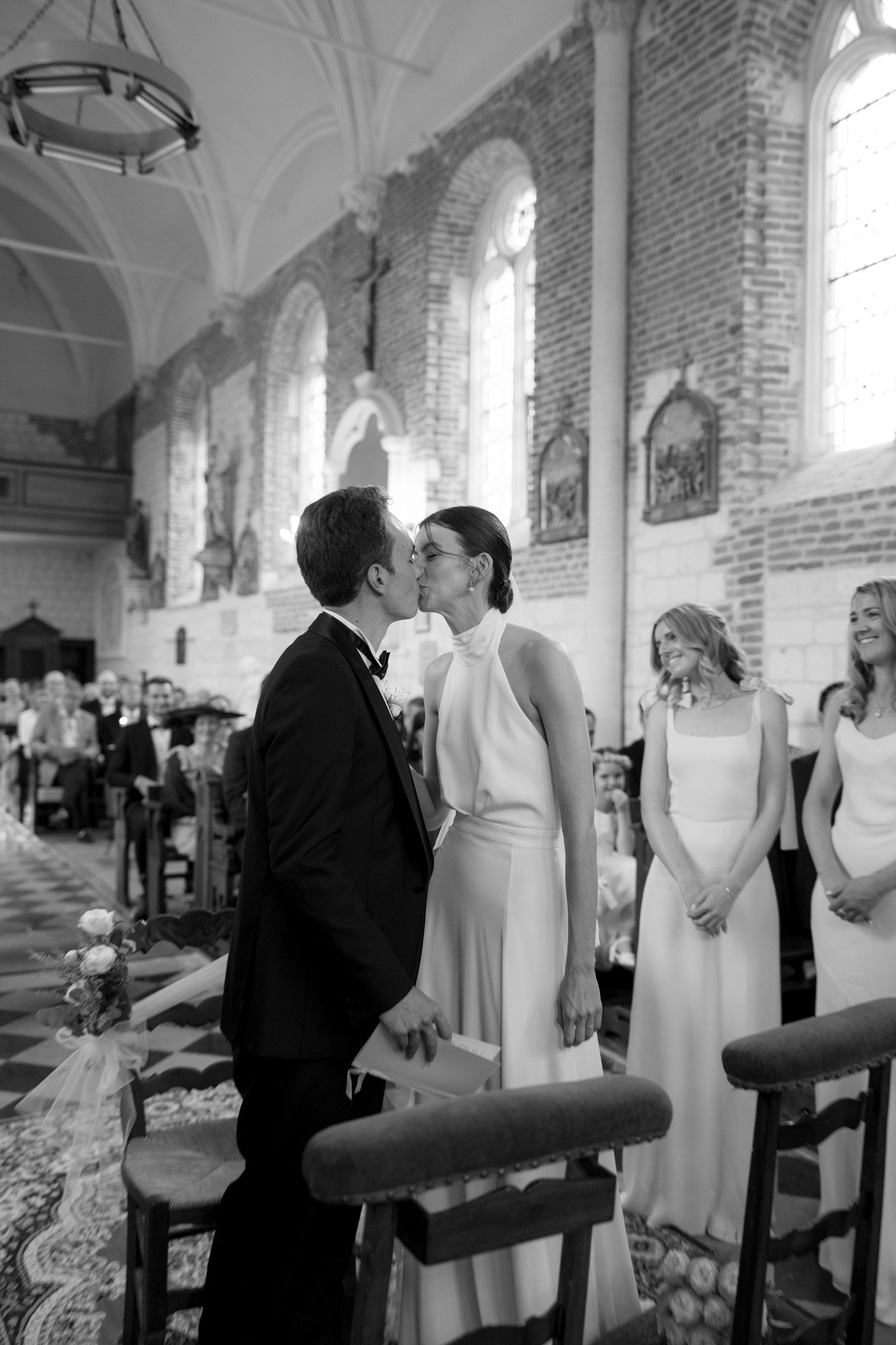 A bride and groom kiss inside a church during their wedding ceremony, with guests seated and bridesmaids standing nearby.