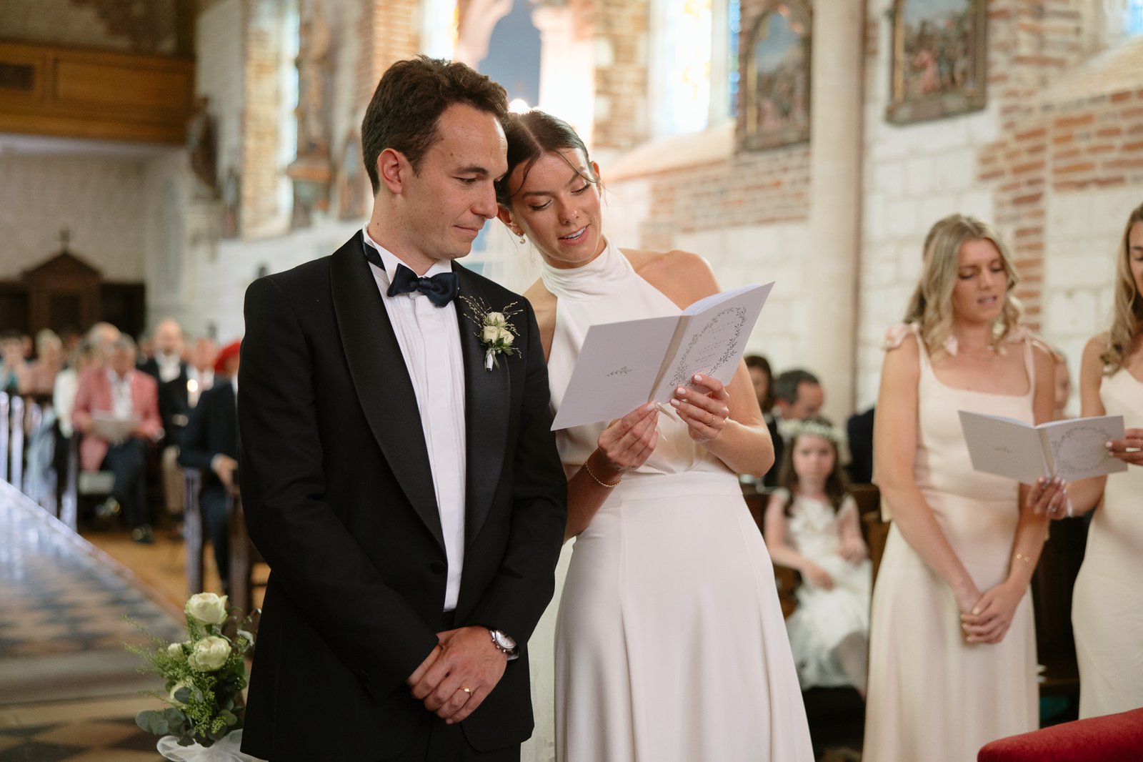 A bride and groom stand together at the altar, reading from a card during their wedding ceremony in a church, with guests seated in the background.