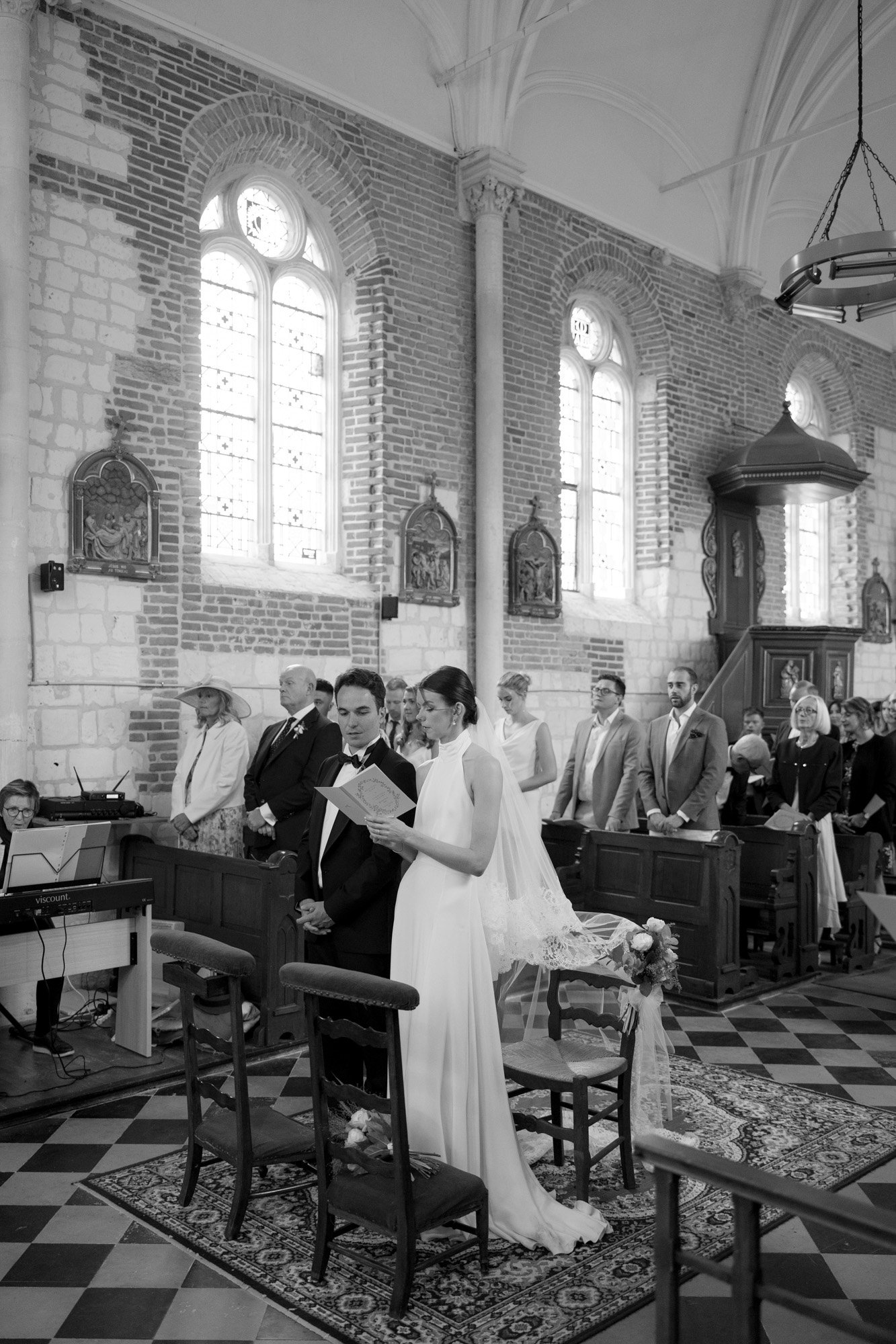 A bride and groom stand at the altar reading from a booklet in a church, with guests seated and standing behind them during a wedding ceremony.