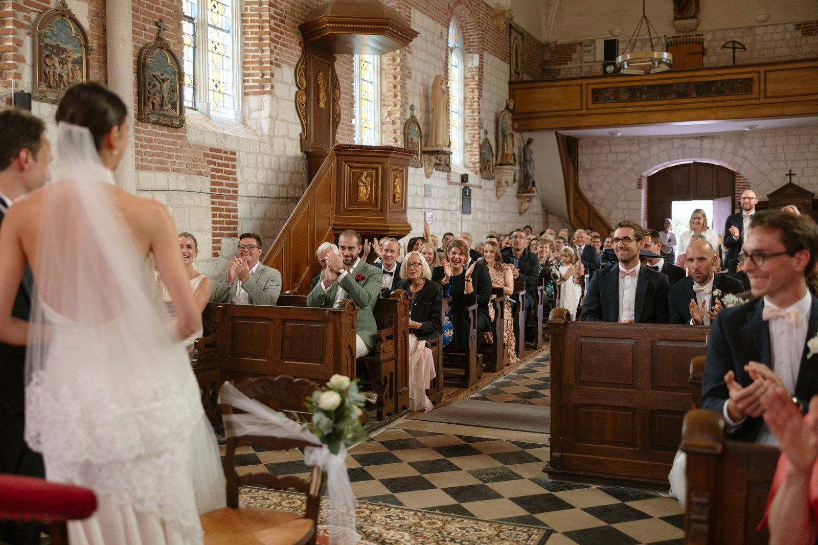 A bride and groom stand at the altar in a church as guests sit in pews, applauding and smiling during a wedding ceremony.