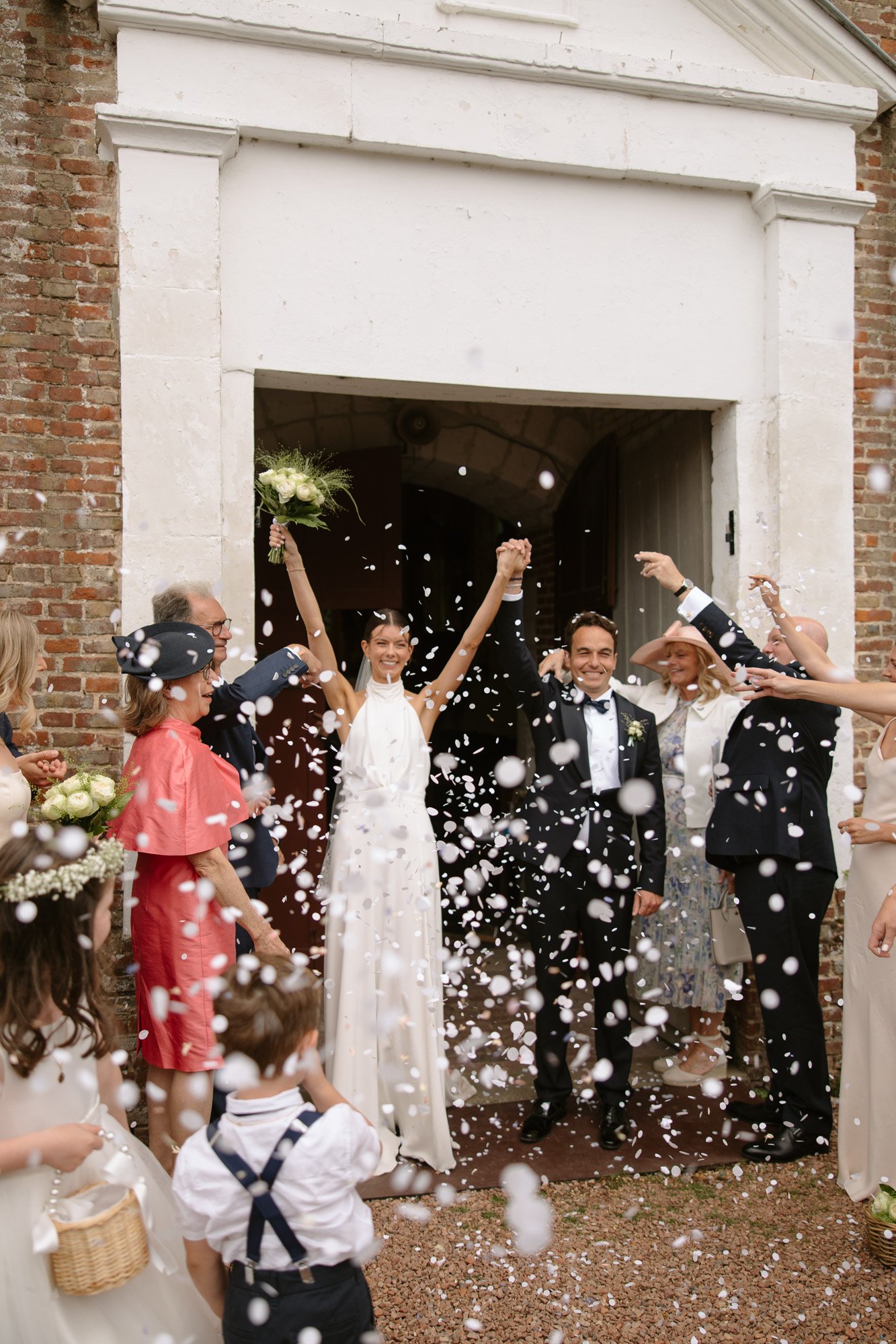 A newlywed couple exits a building as guests celebrate by throwing flower petals; the bride lifts her bouquet and hand in the air, both smiling. Captured by Northern France wedding photographer.