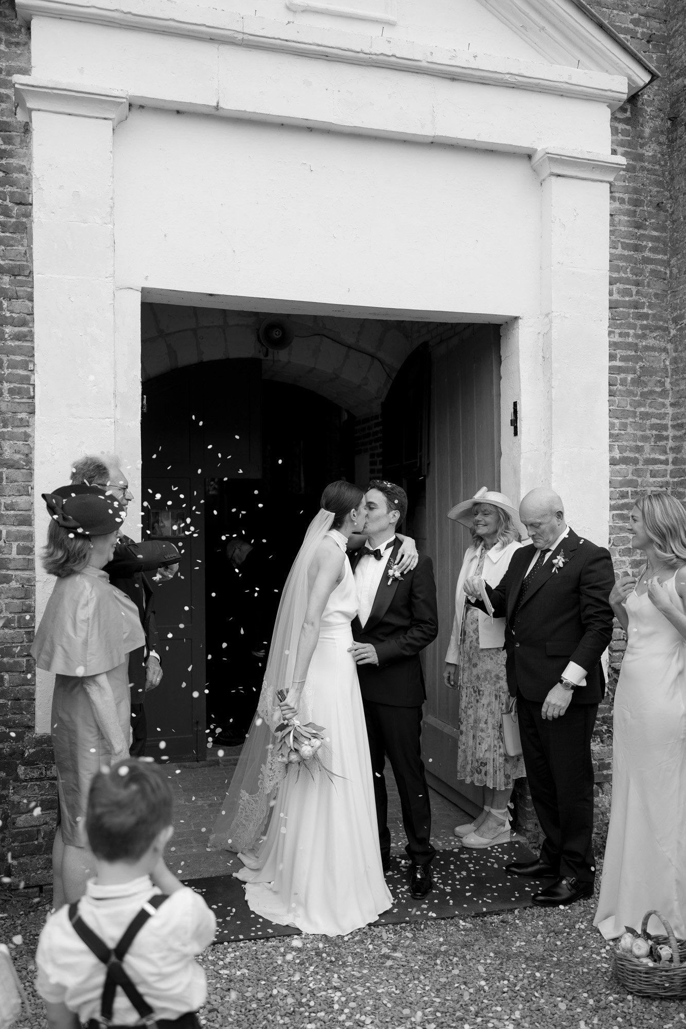 A bride and groom kiss at the church entrance as guests surround them and flower petals are thrown in celebration. Captured by Northern France wedding photographer.