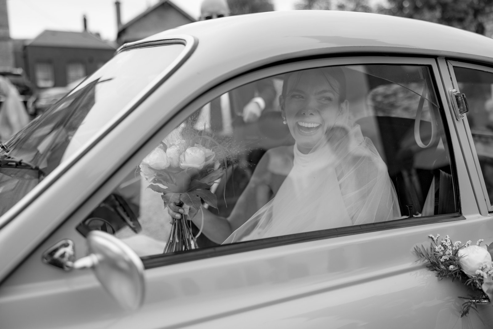 A bride holding a bouquet smiles and looks out the window from inside a car, with wedding decorations visible on the vehicle. Captured by Northern France wedding photographer.