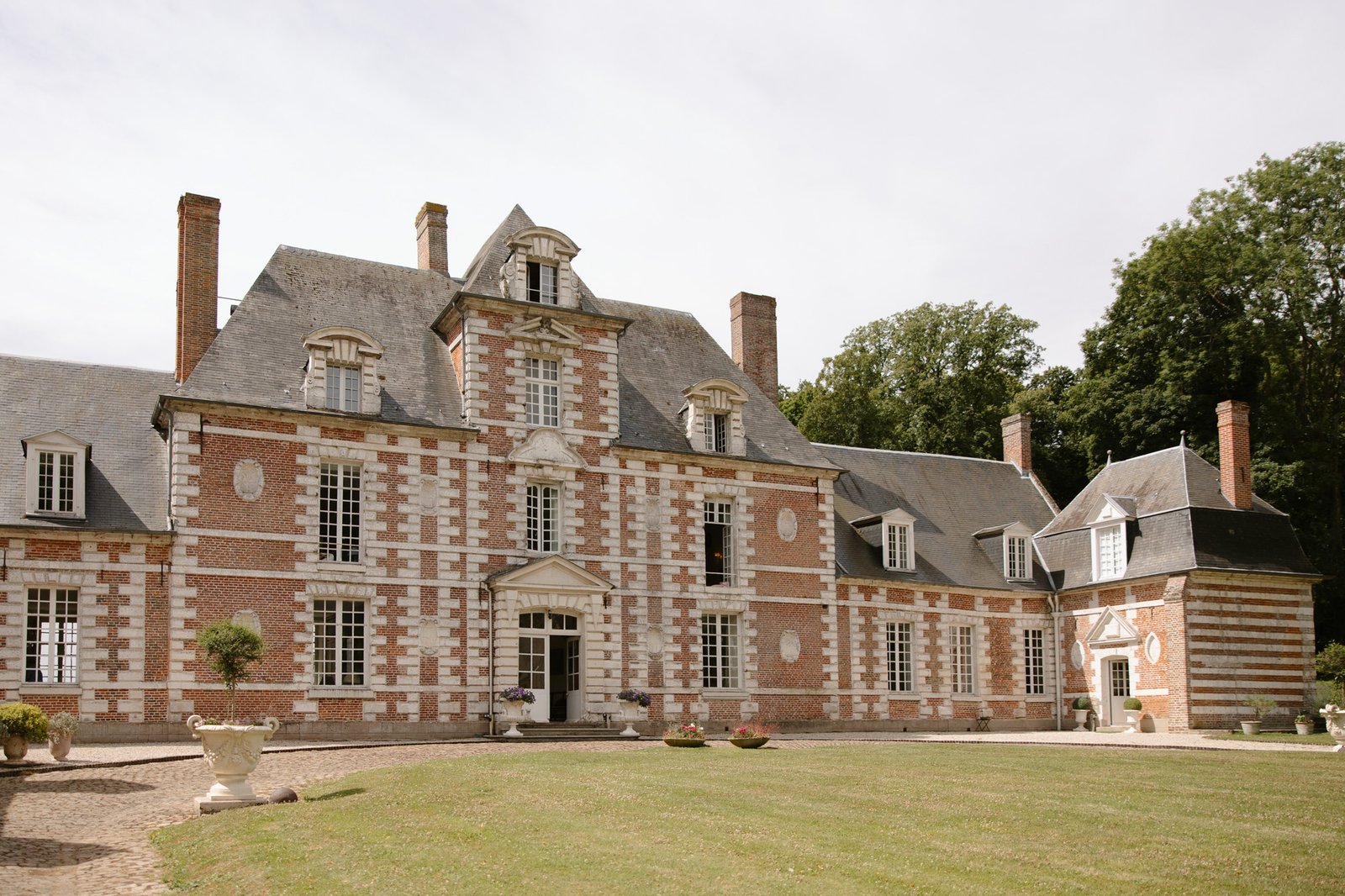 A large, historic brick and stone chateau with tall chimneys and dormer windows, surrounded by a lawn and potted plants. Captured by Northern France wedding photographer.