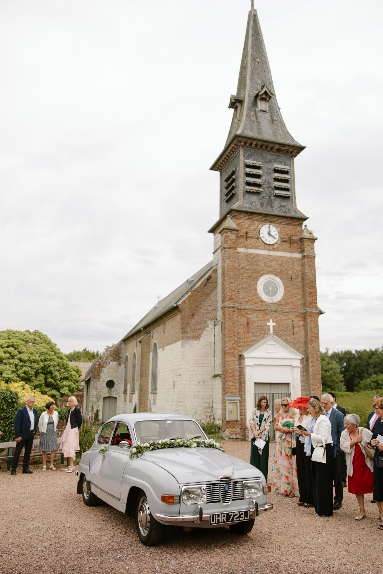 A vintage silver car decorated with flowers is parked in front of a brick church as a group of people dressed formally stand nearby.