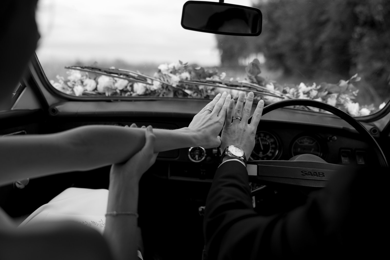 A couple in a car holds hands over the steering wheel, with a floral arrangement on the dashboard; the image is in black and white.