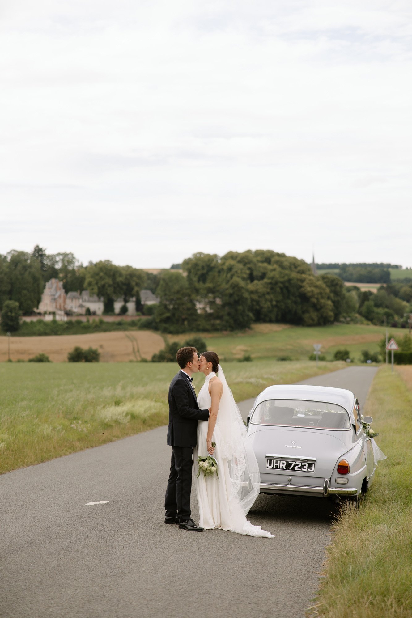 A bride and groom stand close together on a rural road beside a vintage car, with fields and trees in the background. Captured by Northern France wedding photographer.