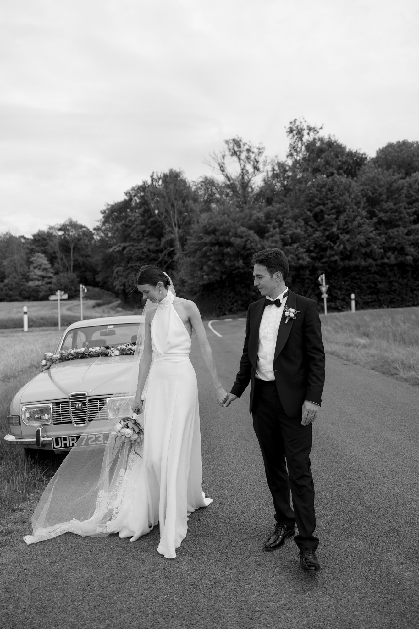 A bride and groom stand on a rural road near a vintage car decorated for a wedding, holding hands and looking down in a black and white photo. Captured by Northern France wedding photographer.