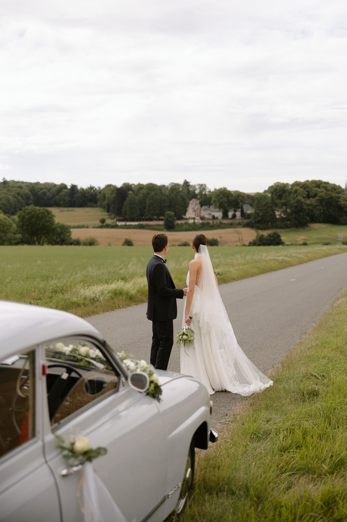 A bride and groom stand together on a rural roadside, facing a scenic landscape, with a decorated vintage car parked nearby.