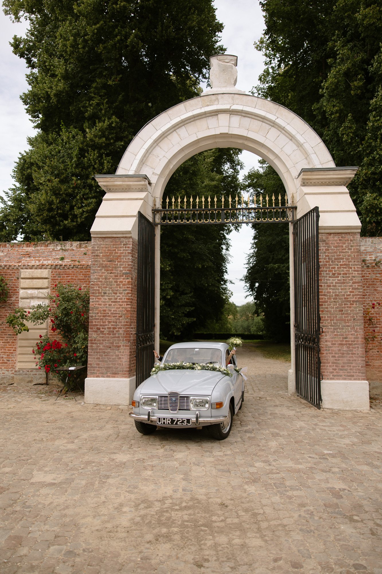 A vintage silver car with a flower garland on the hood is parked under an open brick and stone archway with wrought iron gates.