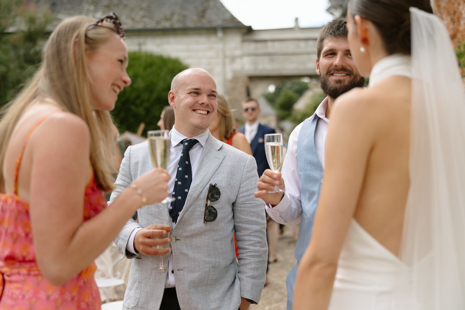 Four people dressed in semi-formal attire stand outdoors, holding champagne glasses and conversing at what appears to be a wedding or celebration.