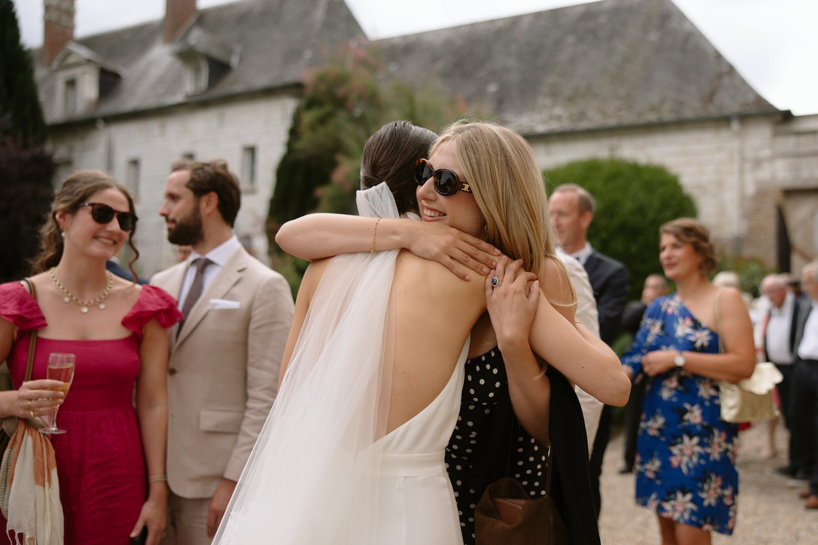 A bride in a white dress hugs a woman in sunglasses at an outdoor gathering, with other guests mingling in the background.