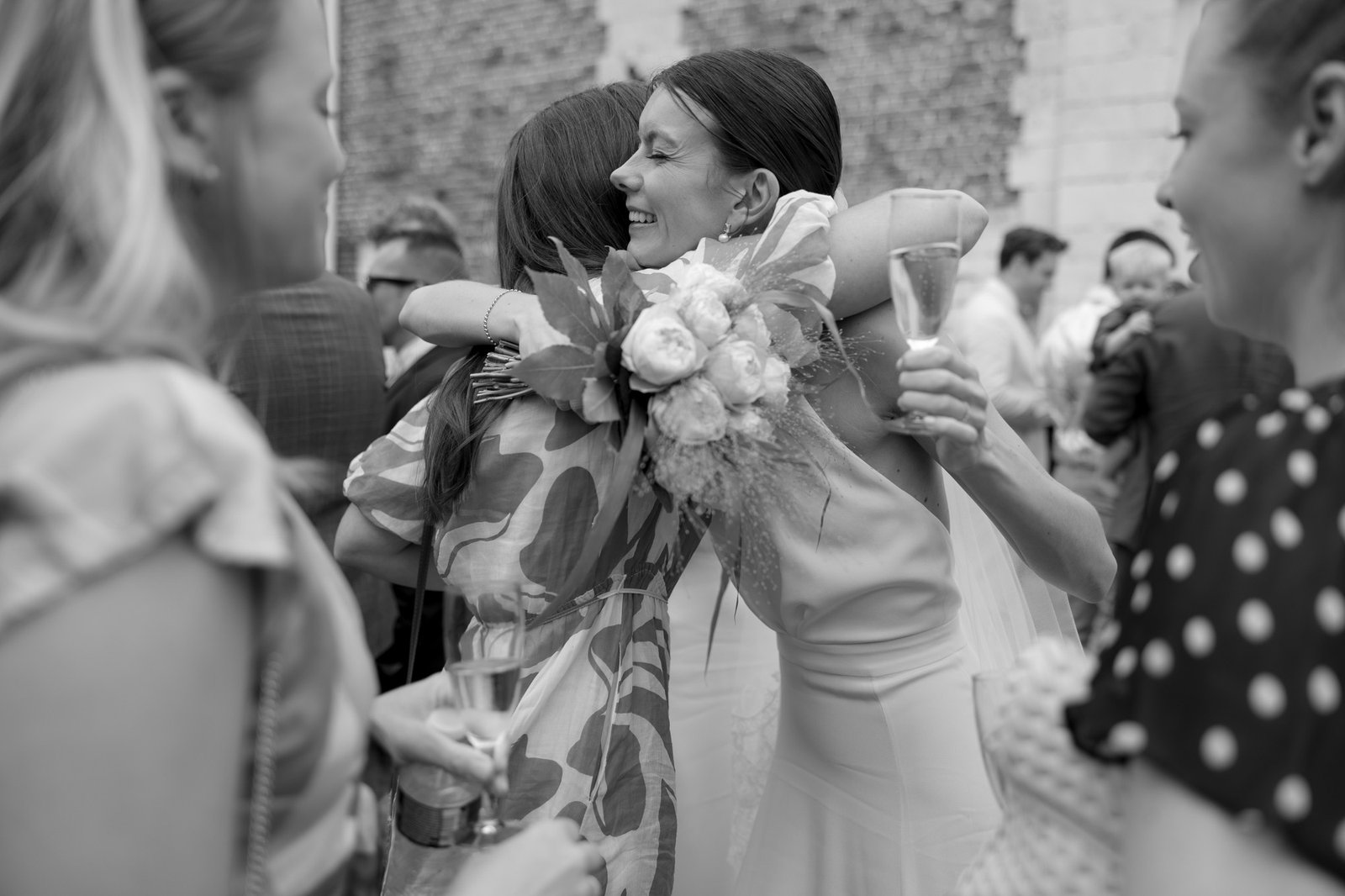 A bride holding a bouquet and a glass of champagne hugs a guest at an outdoor wedding celebration, surrounded by other attendees.
