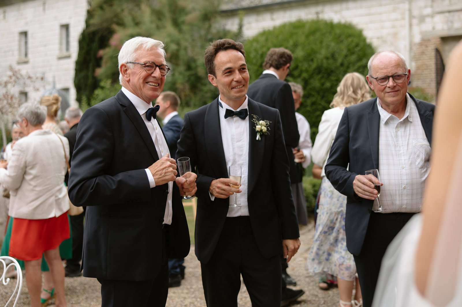 Three men in suits, two wearing bow ties, hold drinks and smile at an outdoor event with other guests in the background.