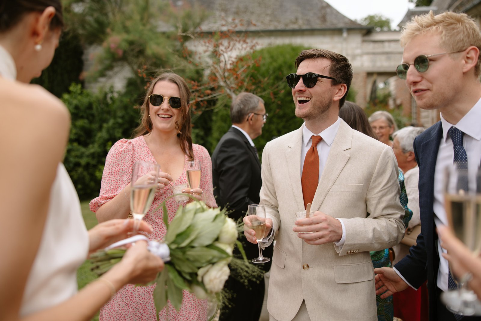 A group of people in formal attire stand outdoors, holding drinks and conversing at an event. One person holds a bouquet of flowers. Captured by Northern France wedding photographer.