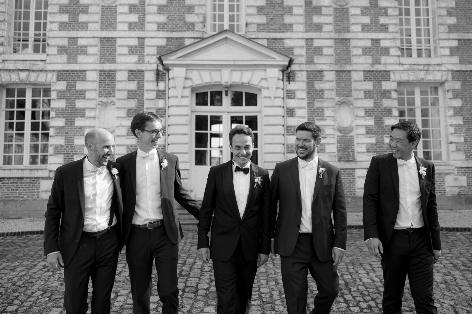 Five men in suits, including one in a bow tie, walk together and smile in front of a large brick building with tall windows. Captured by Northern France wedding photographer.