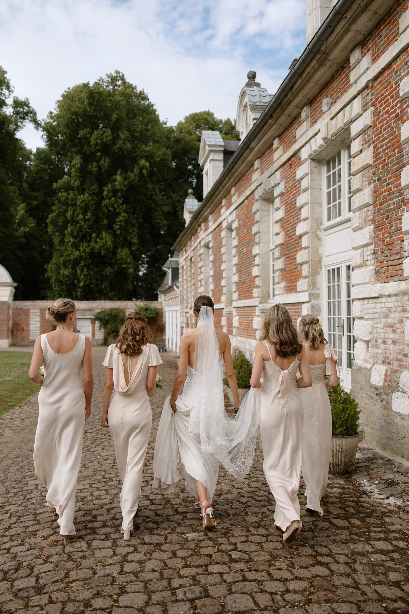 Five women in light dresses, including a bride in a veil, walk on a cobblestone path beside a brick building with white trim and large windows.