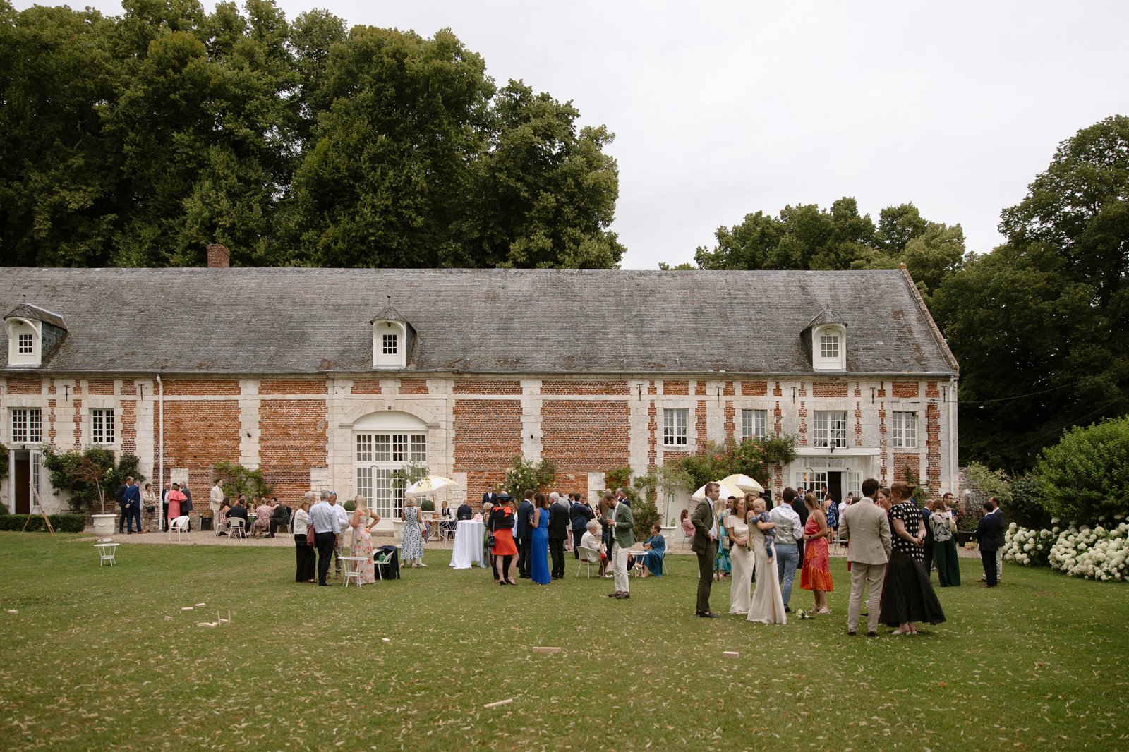 People gather and socialize on the lawn in front of a large brick estate with white trim and a slate roof, surrounded by trees.