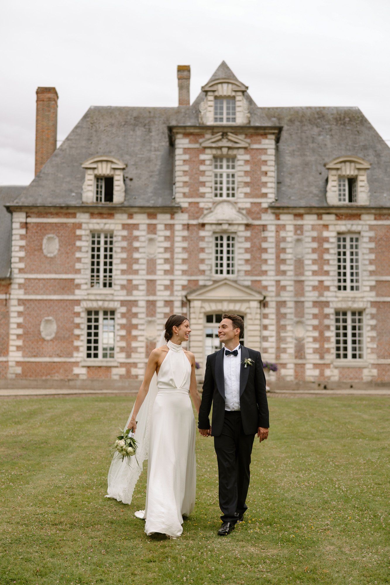 A bride and groom walk hand in hand on a lawn in front of a large brick building with white trim and multiple windows. Captured by Northern France wedding photographer.