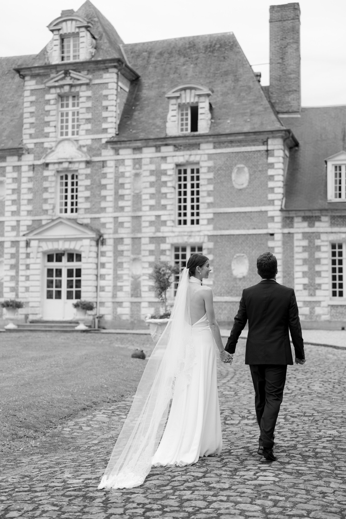 A bride and groom holding hands walk toward a large, historic building with brick and stone features. Captured by Northern France wedding photographer.
