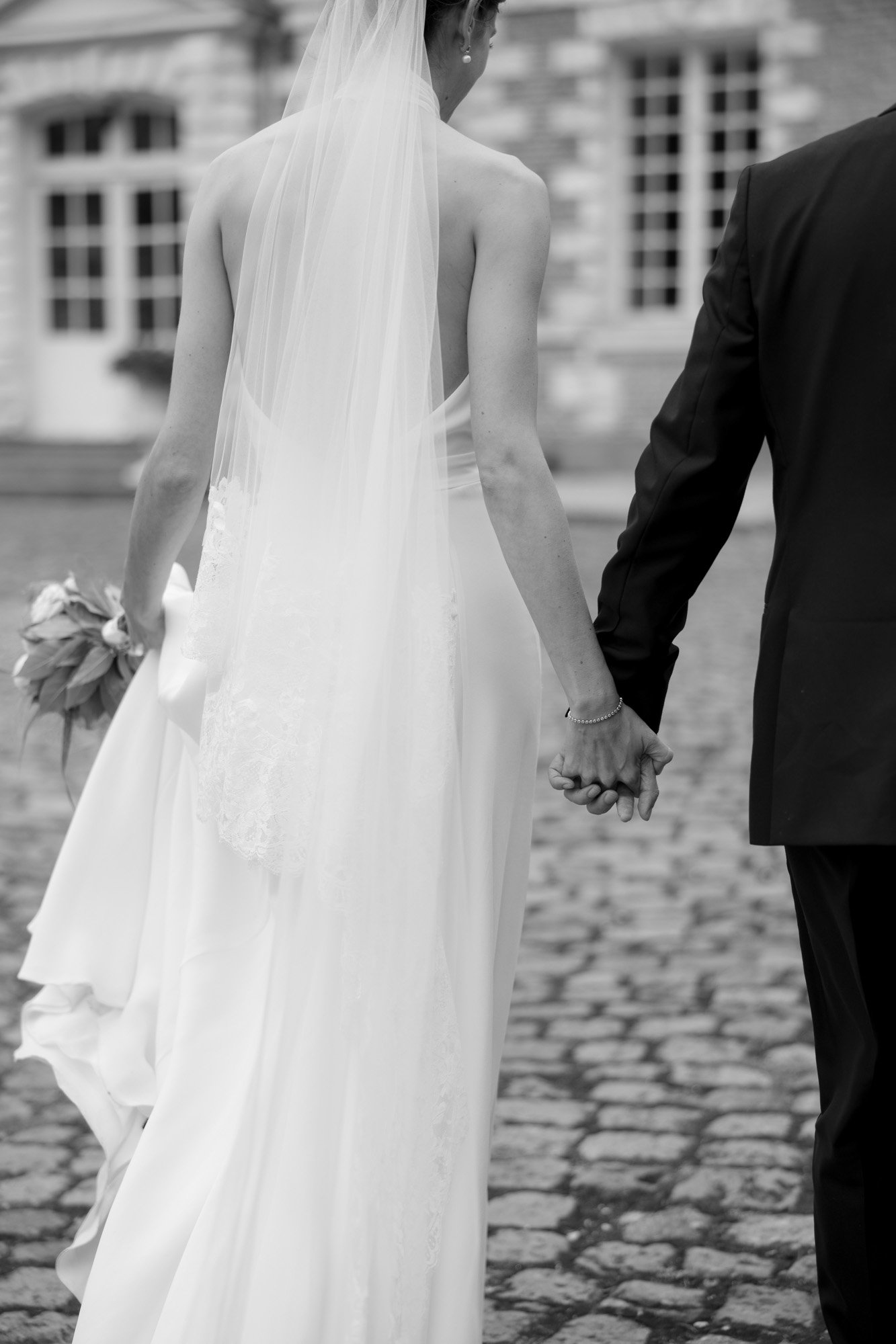 A bride in a white dress and veil holds hands with a person in a dark suit as they walk on a cobblestone path outside a building.