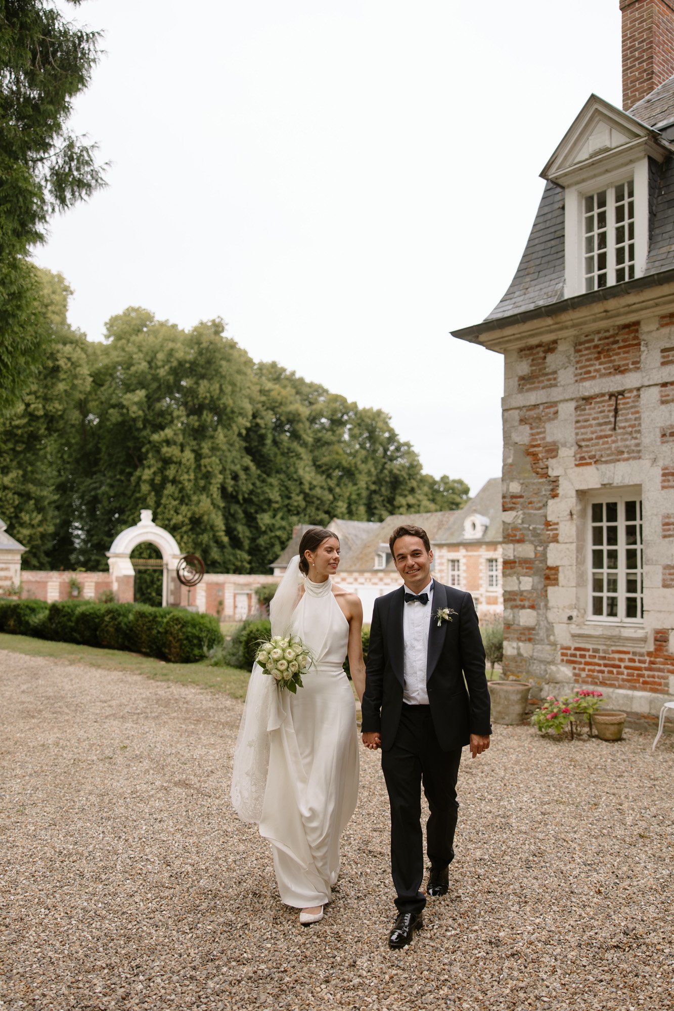 A man and woman in a suit and white dress walking down a gravel path. Captured by Northern France wedding photographer.