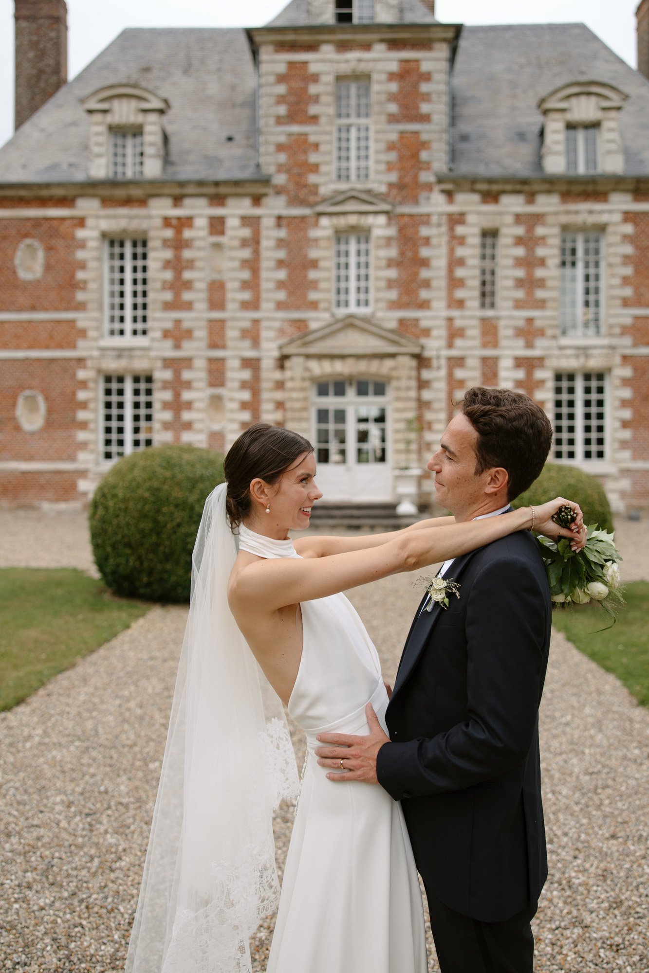 A bride and groom stand facing each other, smiling, outside a large brick mansion with white trim and a gravel pathway. Captured by Northern France wedding photographer.