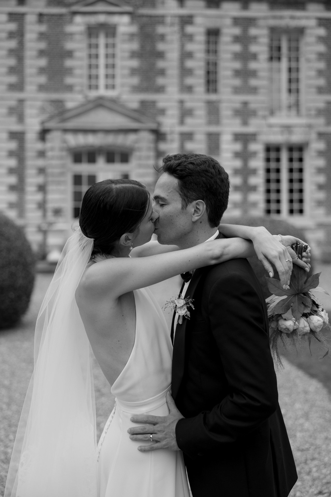 A bride and groom share a kiss outside a large stone building. The bride wears a veil and holds a bouquet; the groom wears a suit.