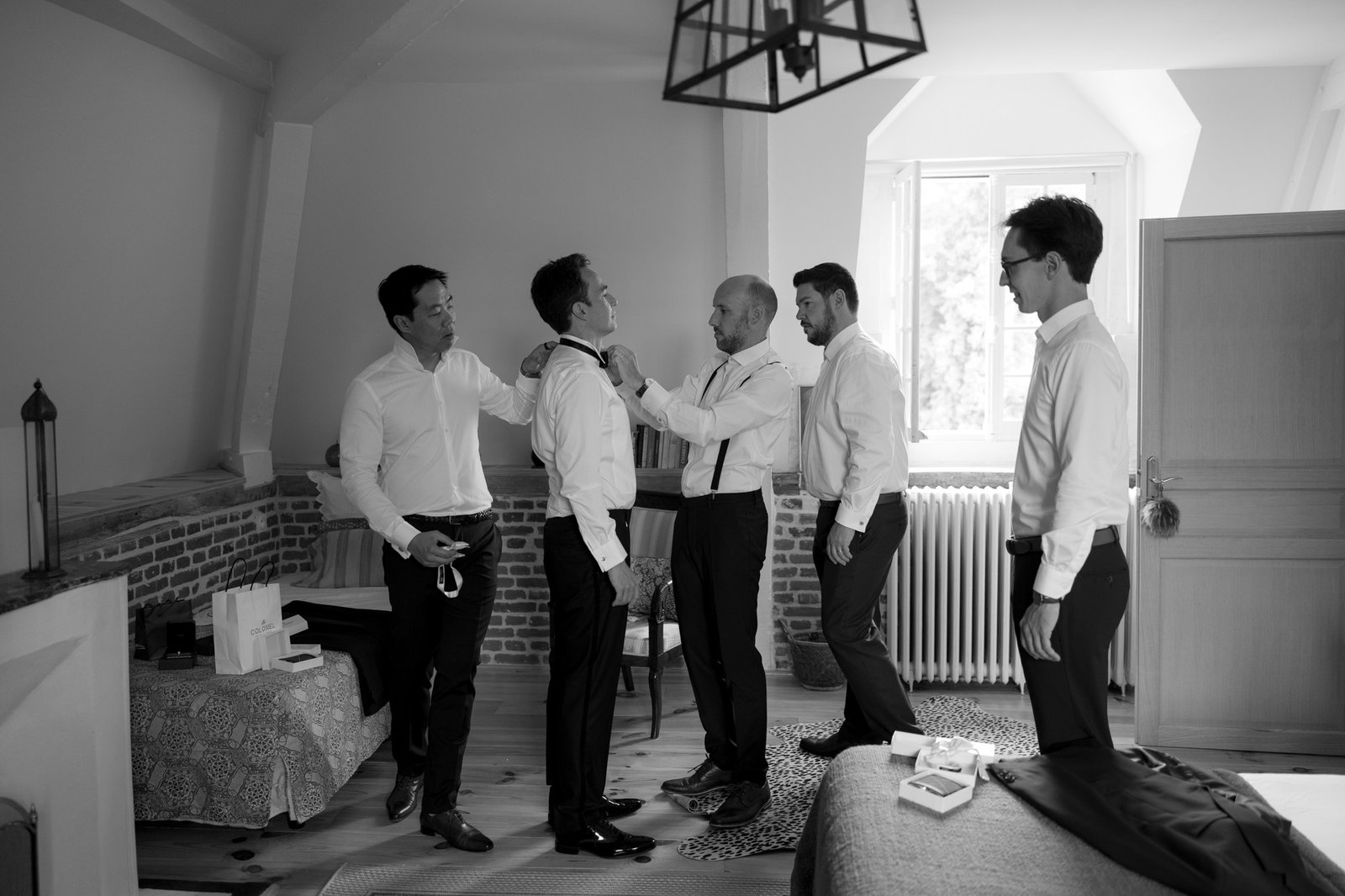 Five men in dress shirts and slacks stand in a room; two are adjusting the tie of another, suggesting preparation for a formal event. Captured by Northern France wedding photographer.