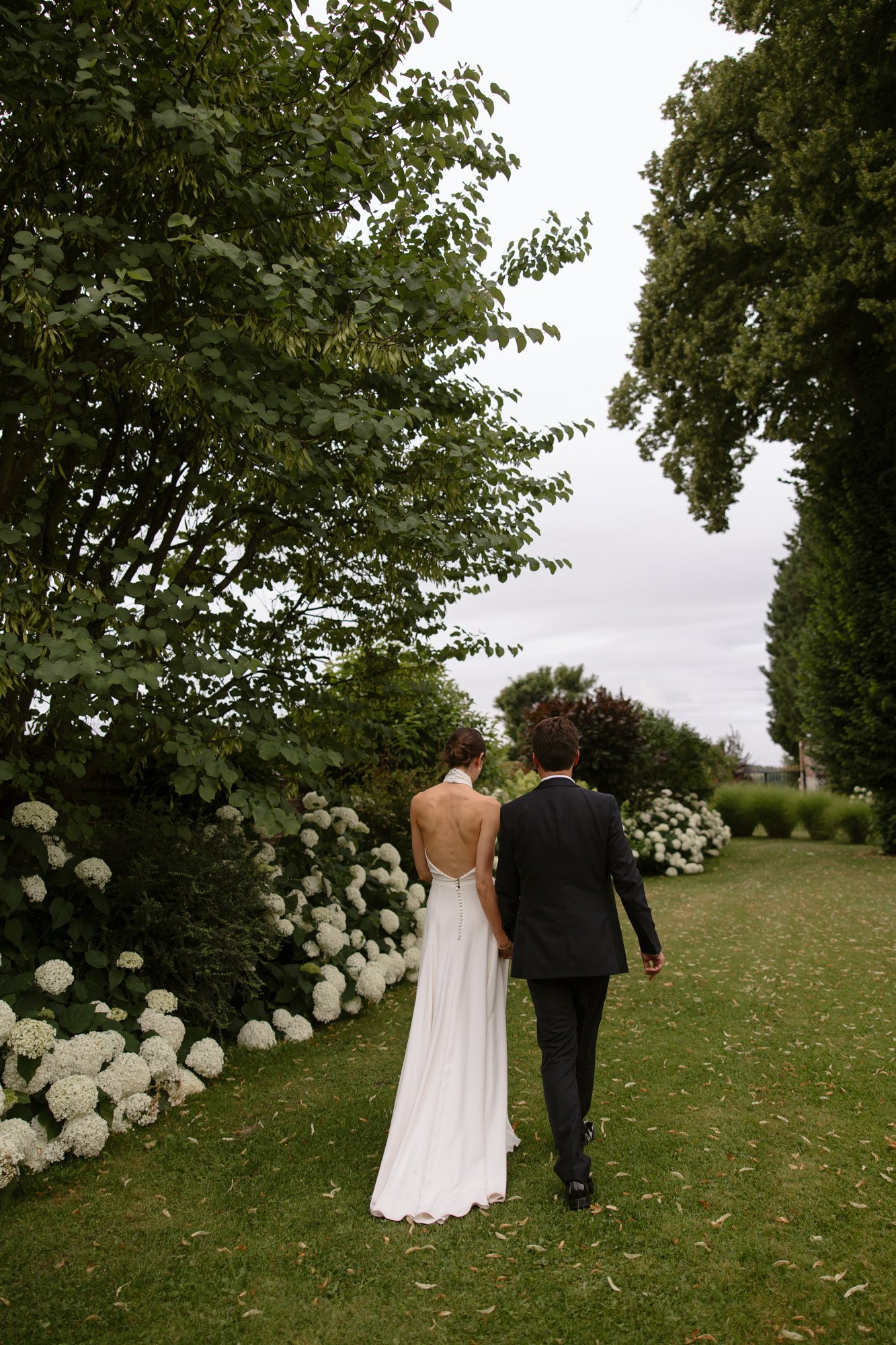 A couple dressed in wedding attire walks hand in hand down a garden path lined with white flowers and greenery. Captured by Northern France wedding photographer.
